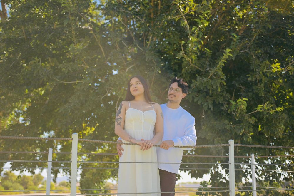 Couple in a loving embrace beside a railing under sunlit trees, captured in a warm outdoor engagement portrait.
