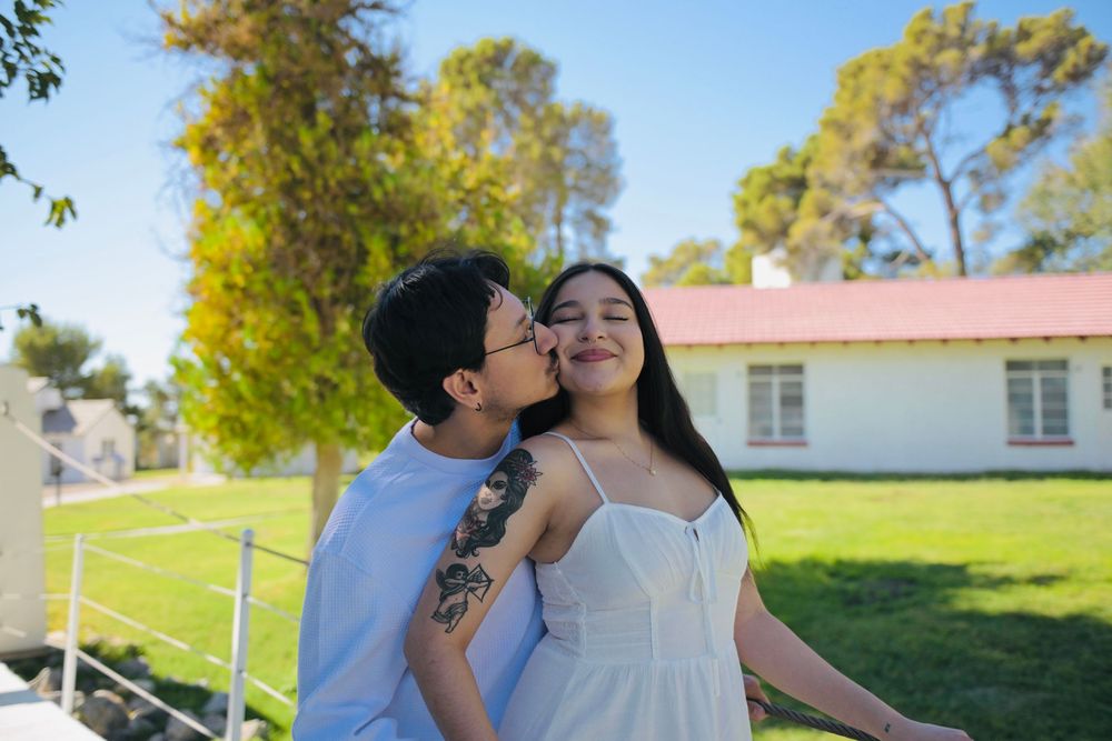Young man in glasses kisses the cheek of a smiling woman in a white sundress with arm tattoos, standing on a sunny green lawn.
