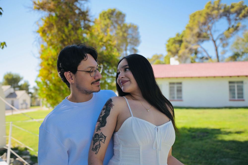 Smiling couple embracing outdoors on a sunny day, the woman in a white dress with arm tattoos, standing on a green lawn near trees.