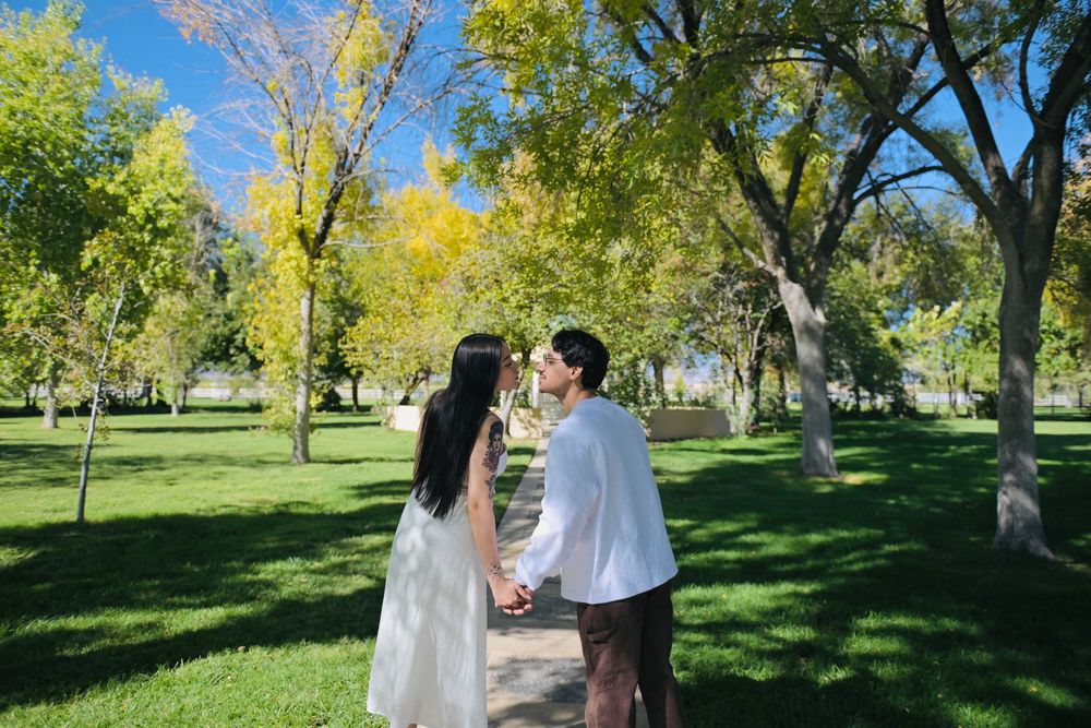 Couple holding hands and walking along a path through a sunny park lined with green and yellow autumn trees.