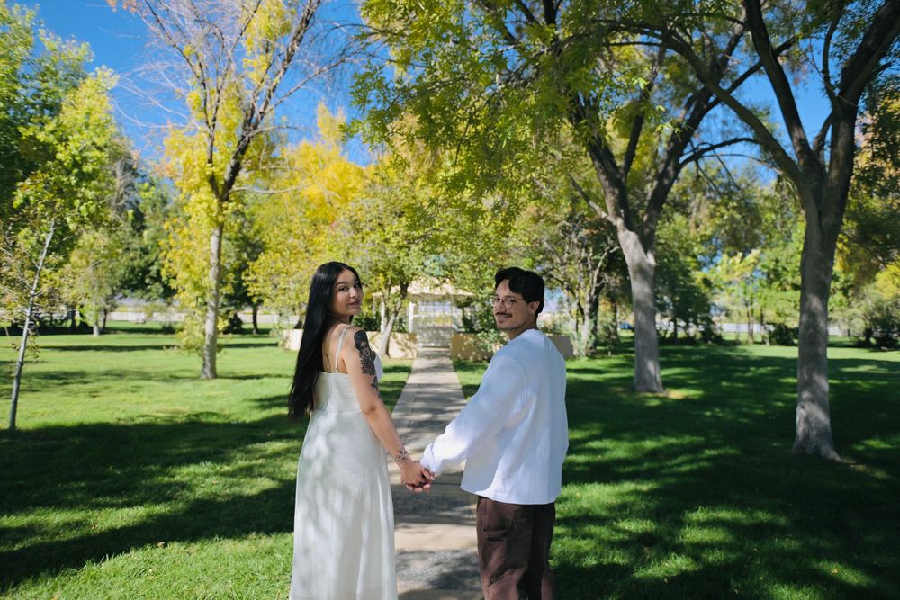 A couple in white clothing holds hands while walking down a sunny tree-lined park path in a romantic engagement photo.