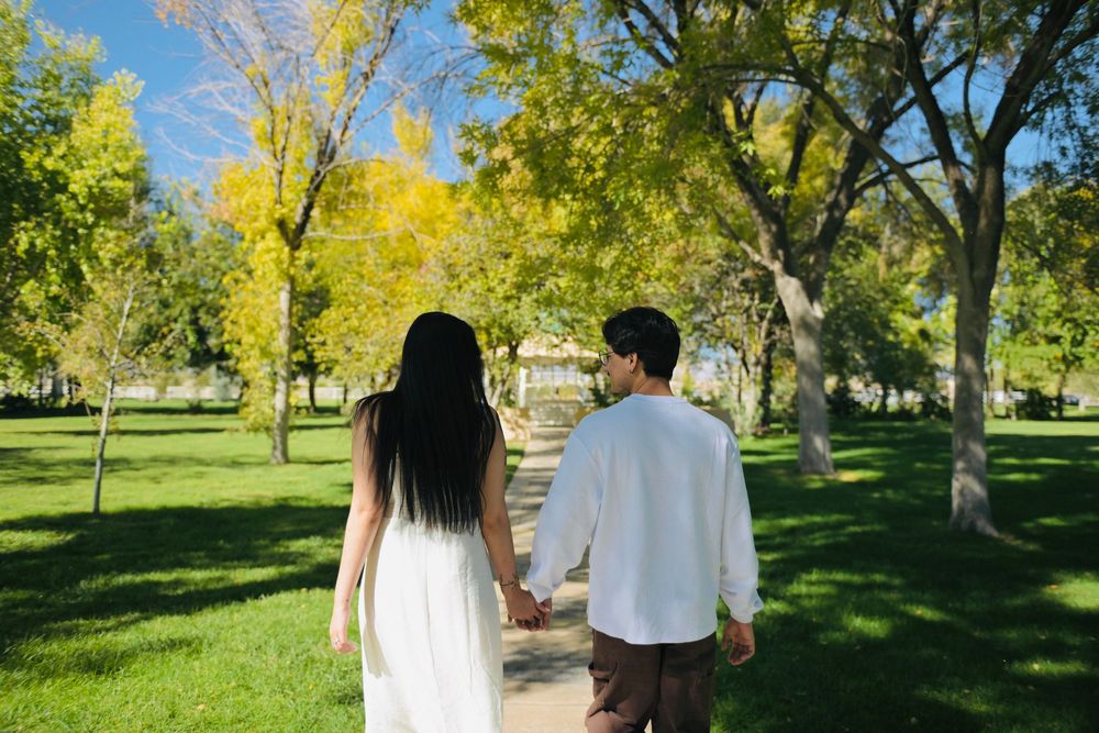Couple holding hands and walking away on a sunlit park path surrounded by green grass and golden autumn trees.