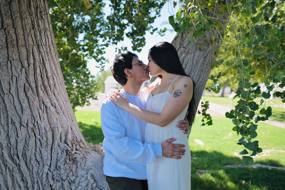 Couple in light clothing embracing and kissing beside a large tree trunk in a sunny green park.