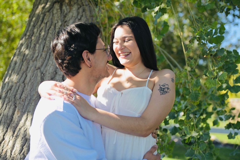 A smiling couple embraces outdoors beside a large tree, the woman in a white sundress laughing as the man in glasses holds her close.
