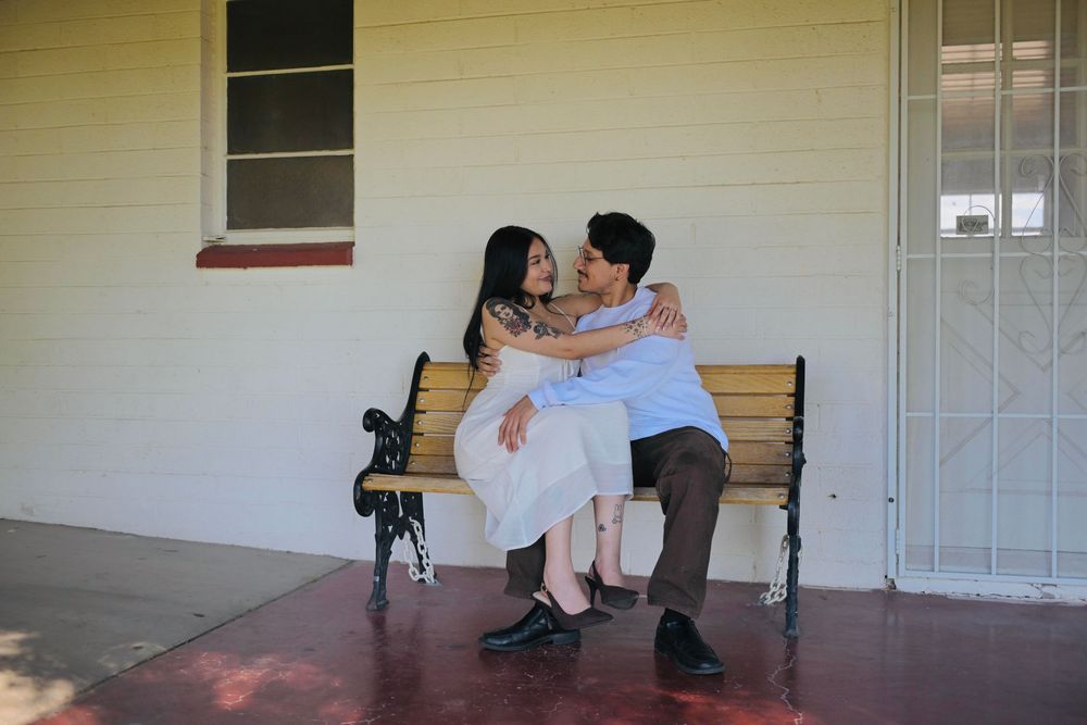 Couple embracing and smiling while seated together on a black wooden park bench in front of a pale yellow wall.