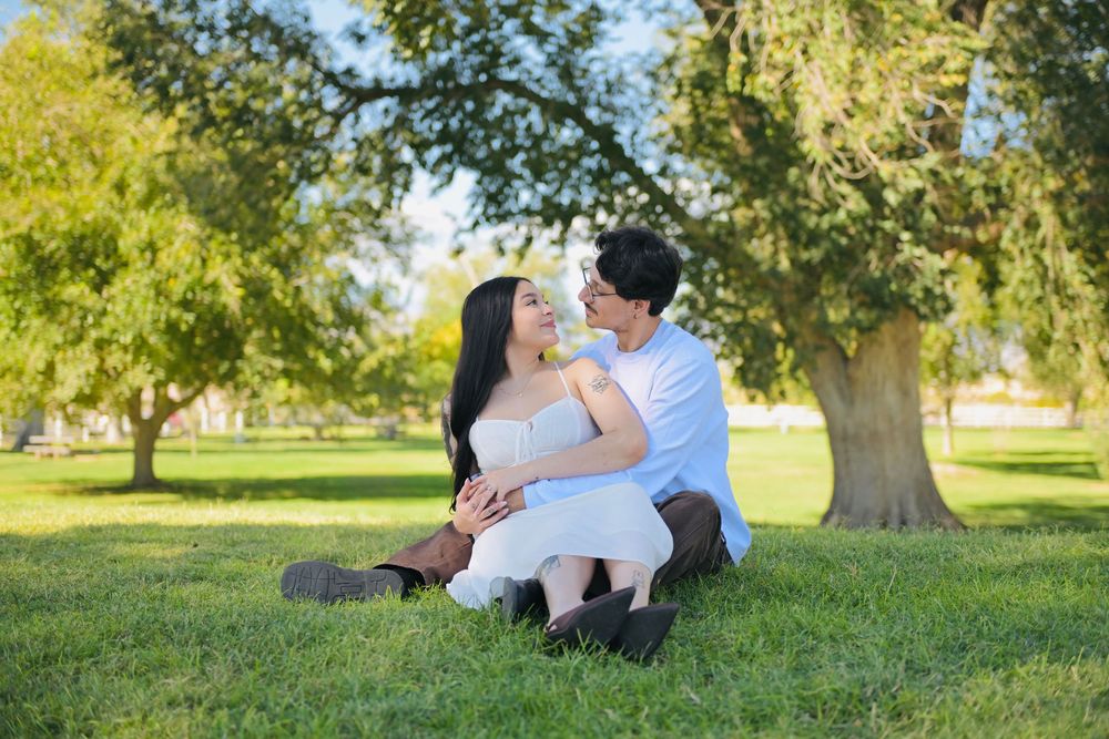 Couple sitting close together on a green grassy lawn under shady trees during a sunny outdoor engagement photo session.