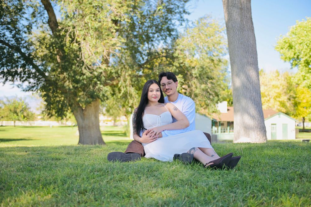 Smiling couple in white outfits embracing on a lush park lawn with tall trees and a small building in the background.