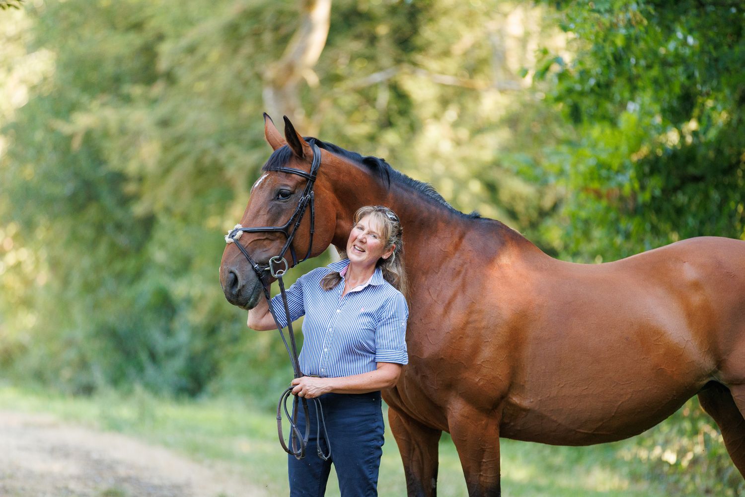 Woman in white lace dress walks alongside a brown horse on a sunlit forest path.