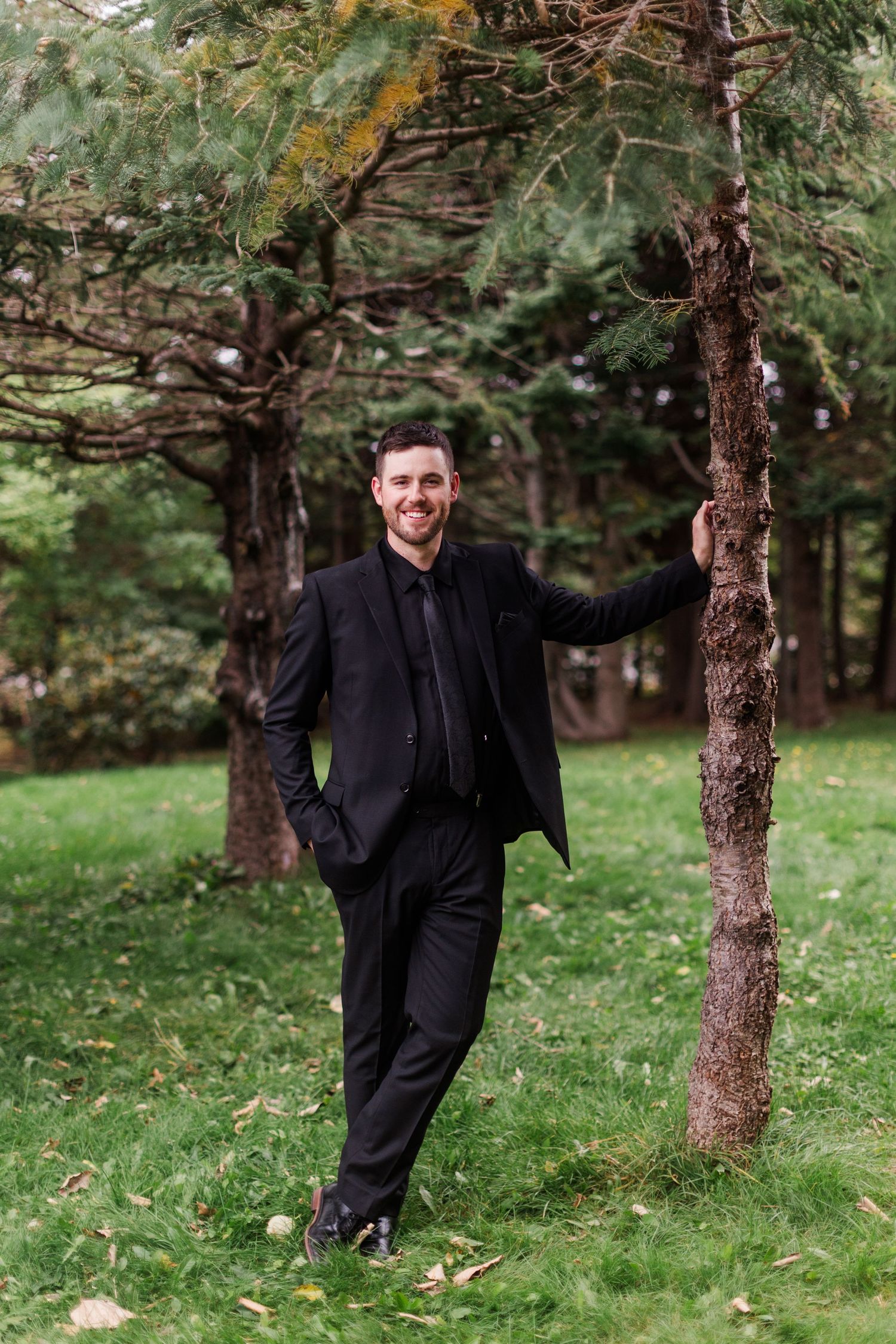Smiling man in a black suit and tie leans against a slender tree trunk on a grassy lawn surrounded by evergreen trees and fallen leaves.