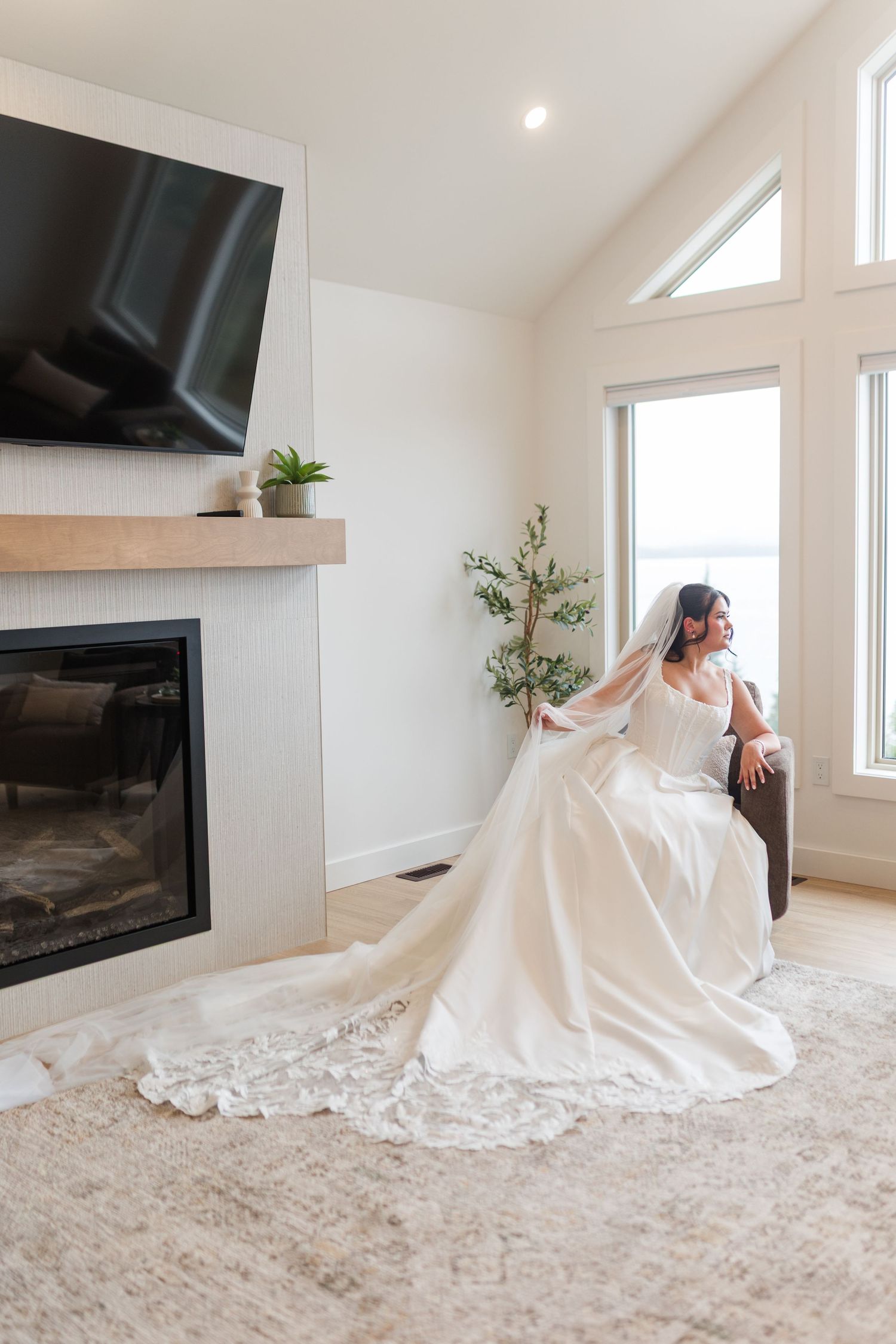 Bride in a white satin wedding gown with lace-trimmed veil sits in a bright living room beside a modern fireplace and large windows.