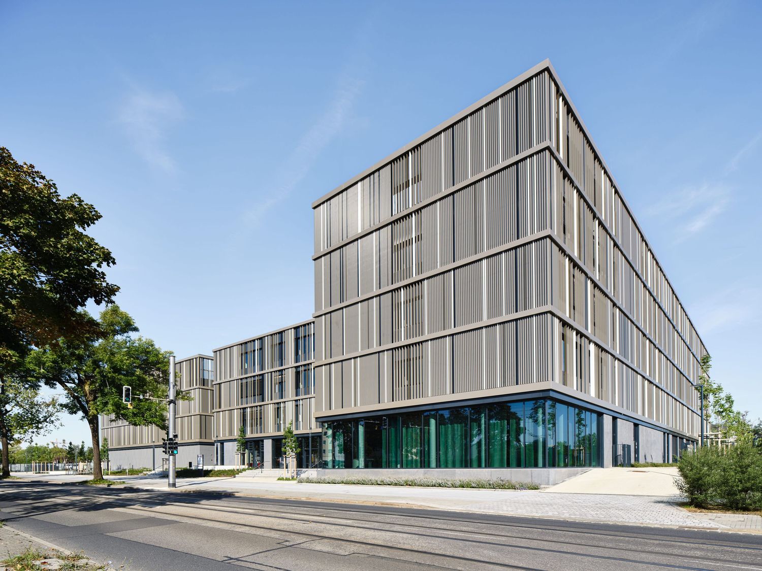 Modern multi-story office building with a vertical slatted metal facade, beside an orange shipping container under a clear blue sky.