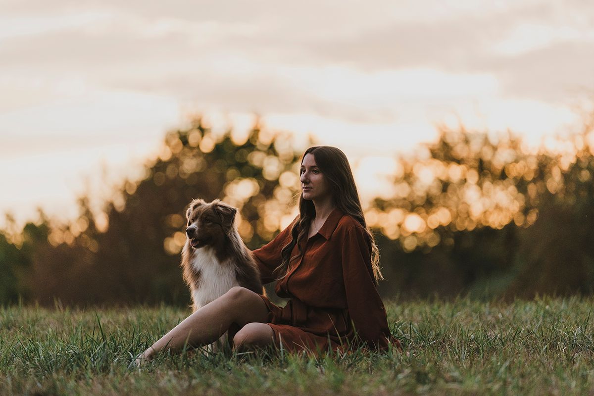 Woman in a rust-colored dress sits in a grassy field with her Australian shepherd as golden sunset light glows behind.