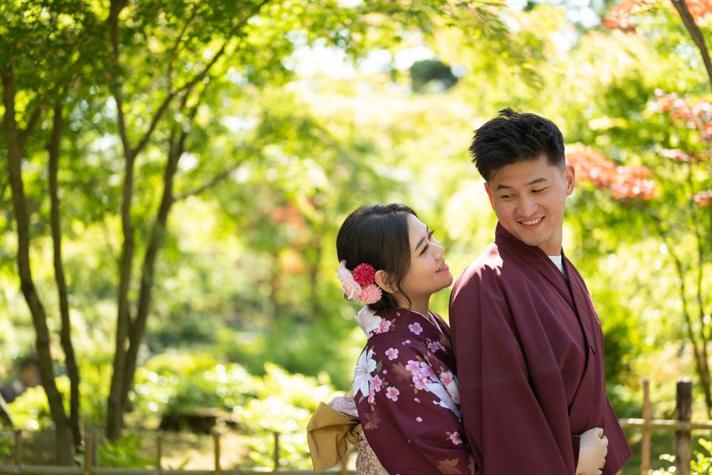 Smiling couple in traditional Japanese yukata, the woman wearing a floral hair accessory, standing together in a lush green garden.