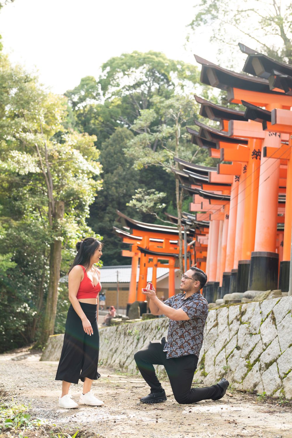 Man kneeling on one knee proposing to a smiling woman in a red top near the vermilion torii gates of Fushimi Inari Shrine in Kyoto.