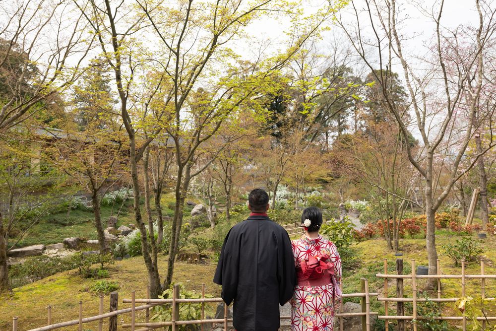 Couple in traditional Japanese kimono stands facing a serene garden with spring trees, moss, and a wooden fence.