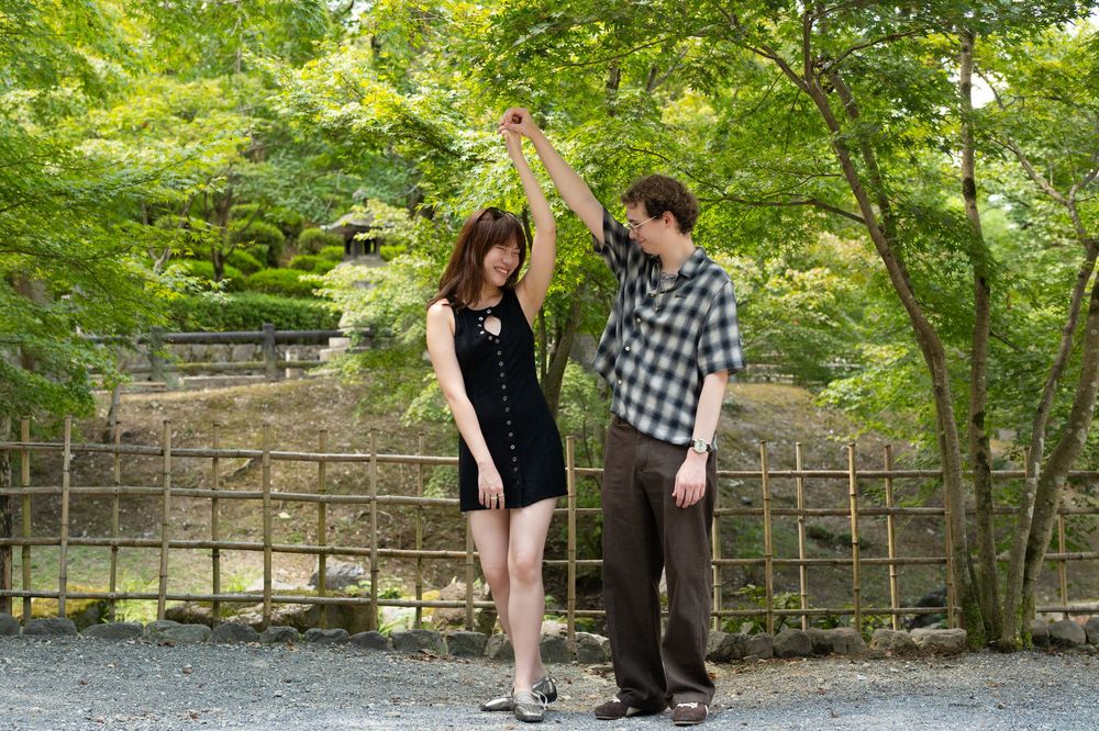A young man in a plaid shirt twirls a smiling woman in a black dress outdoors in a sunny garden with green trees and a wooden fence.