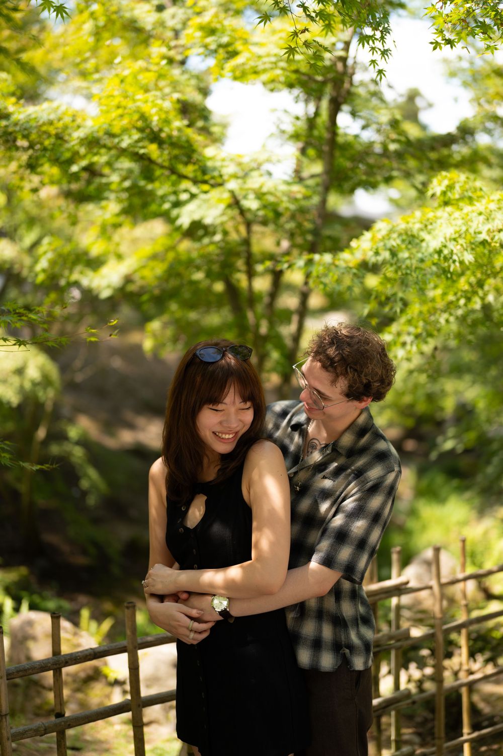 A smiling couple embraces in a sunlit garden, the man hugging the woman from behind beside a wooden railing and green maple trees.