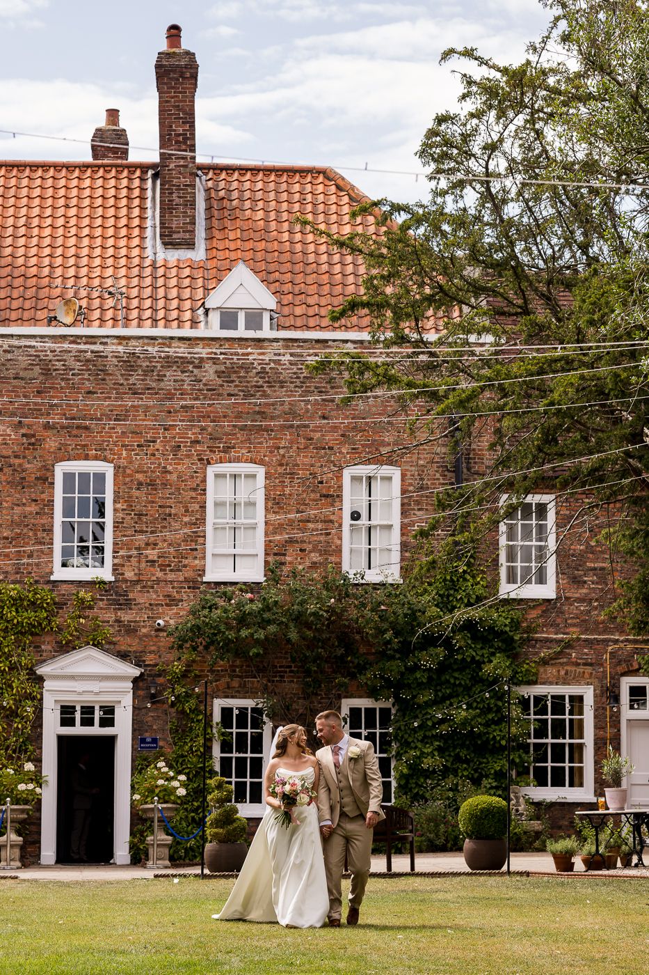 Bride and groom walking hand in hand on lawn before a charming red brick Georgian country house wedding venue.