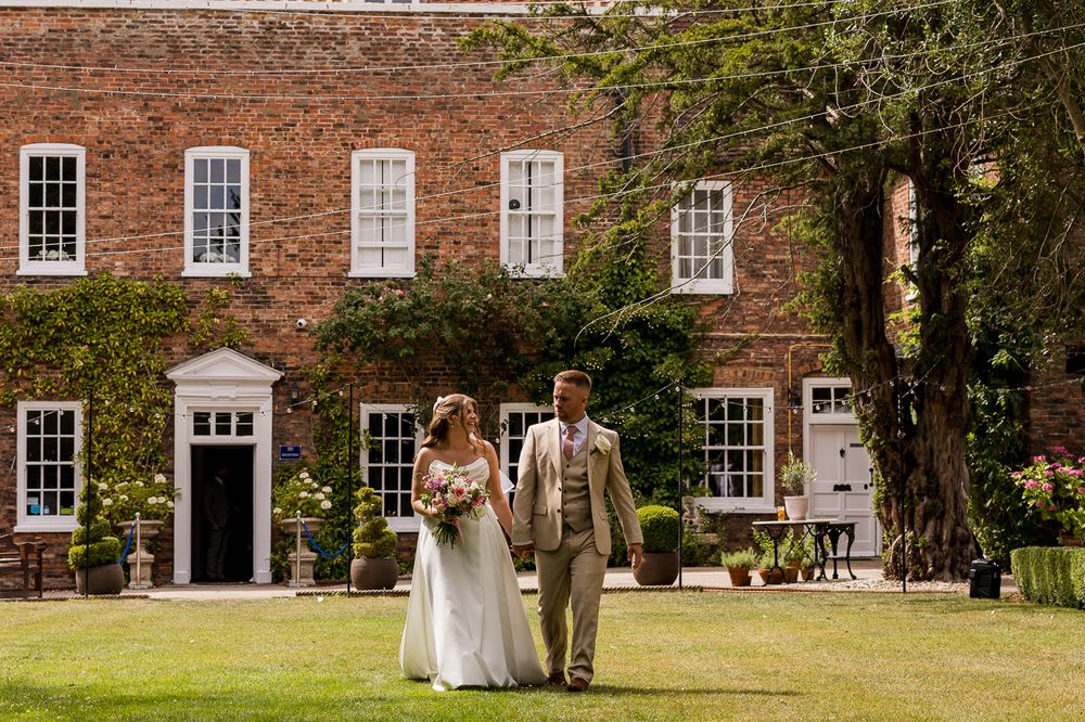 Bride and groom walk hand in hand on green lawn before elegant red brick manor house on sunny wedding day.