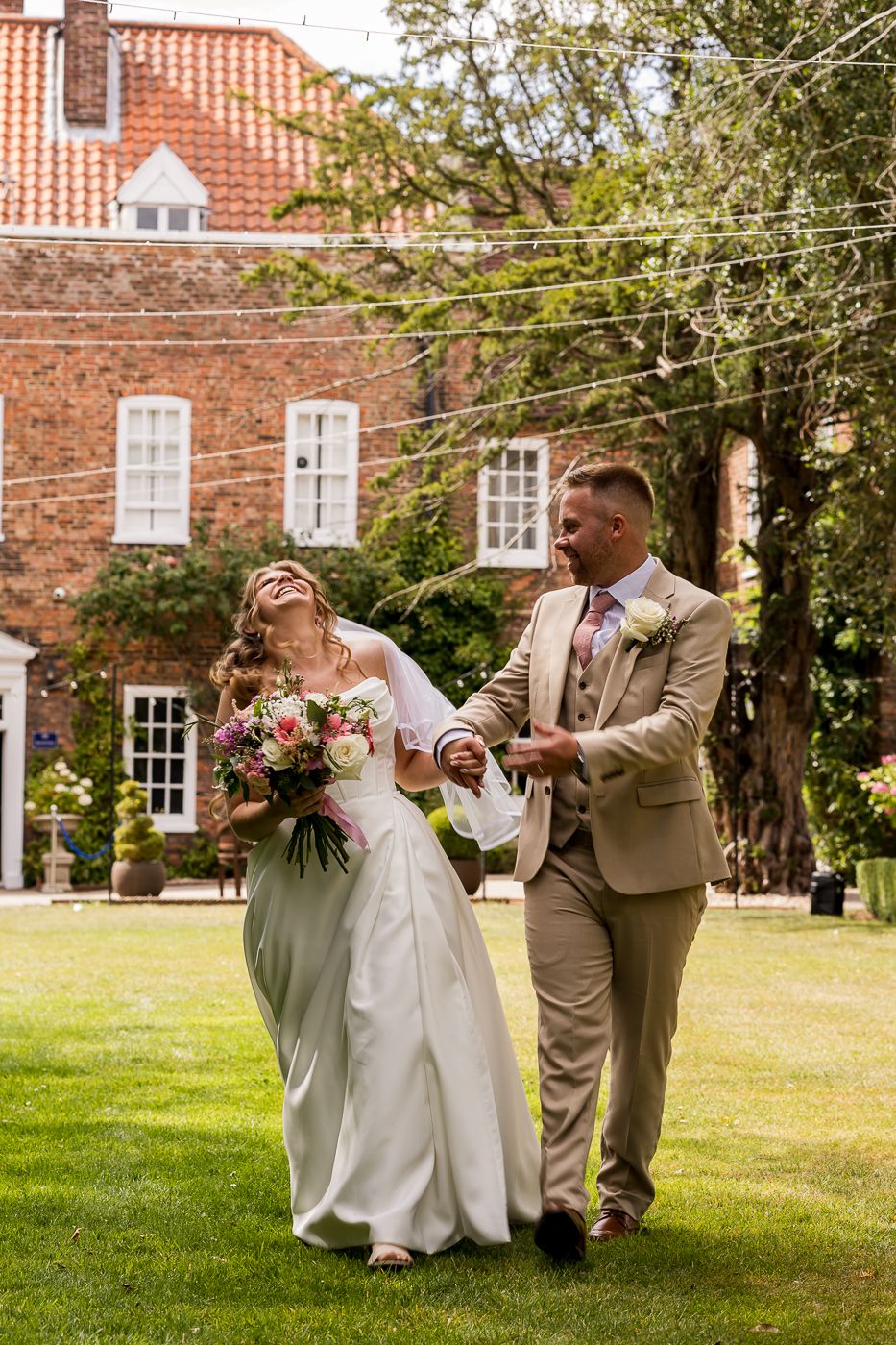 A joyful bride and groom laugh together on a sunny lawn outside a red brick wedding venue.