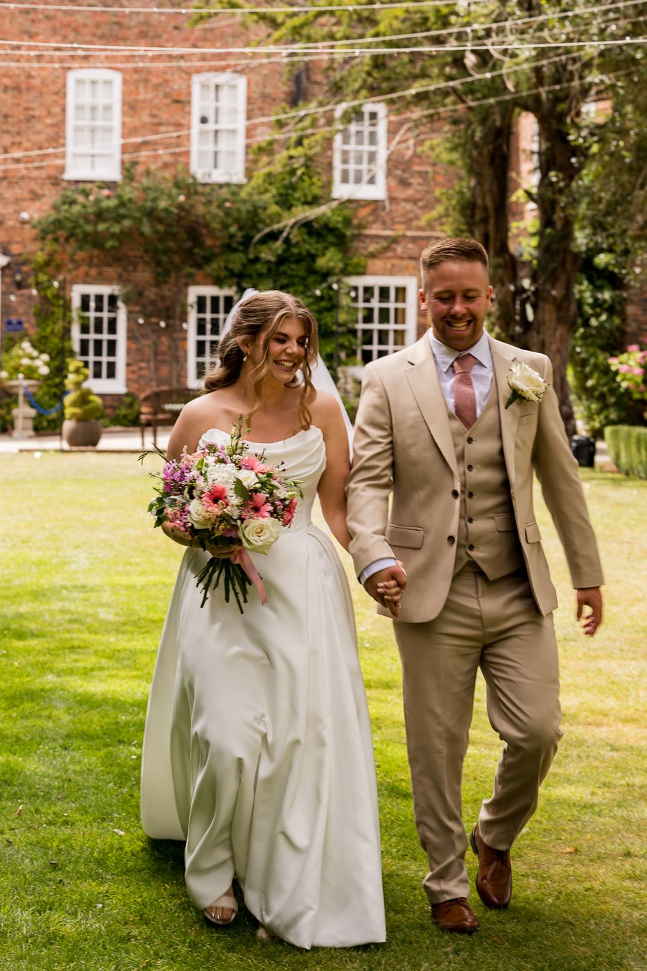 Bride and groom walk hand-in-hand on green lawn, she holds pink bouquet, he wears tan suit, brick manor behind them.