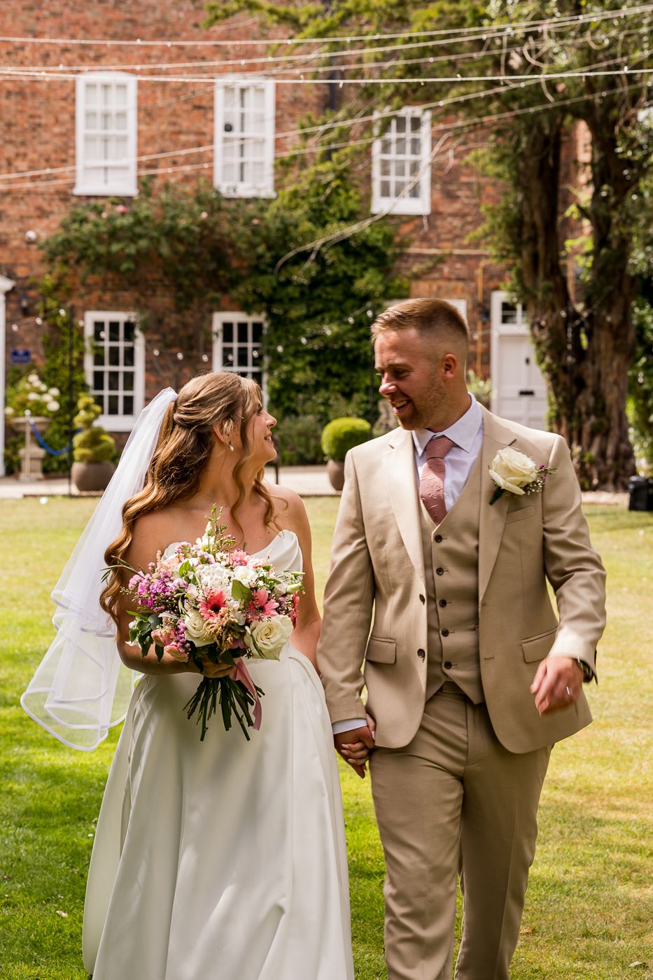 A bride and groom walk hand in hand on a sunny day outside a brick manor, she holds a pink bouquet.