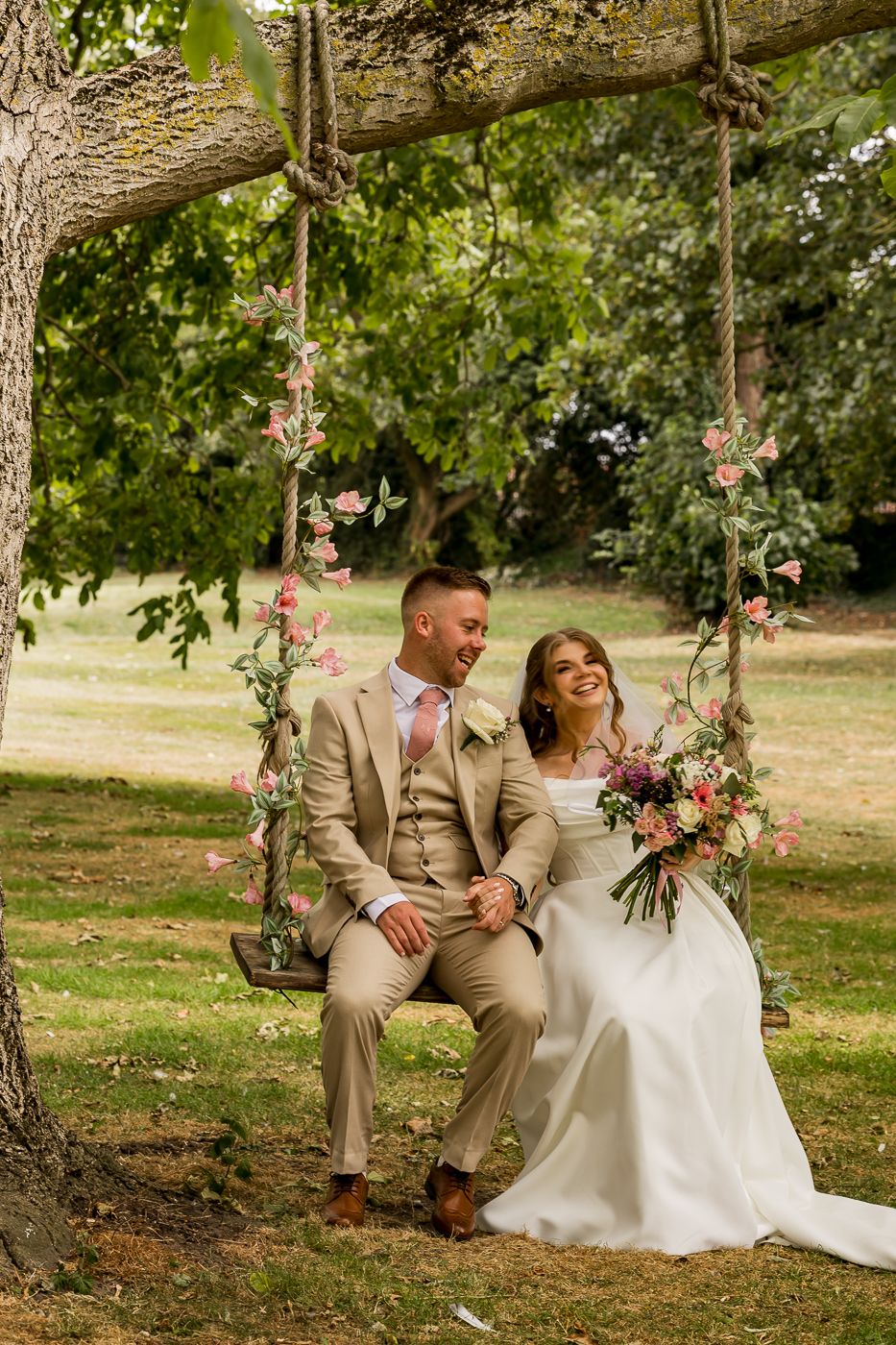 A bride and groom sit together on a floral rope swing under a tree, smiling, as she holds a colorful bouquet.