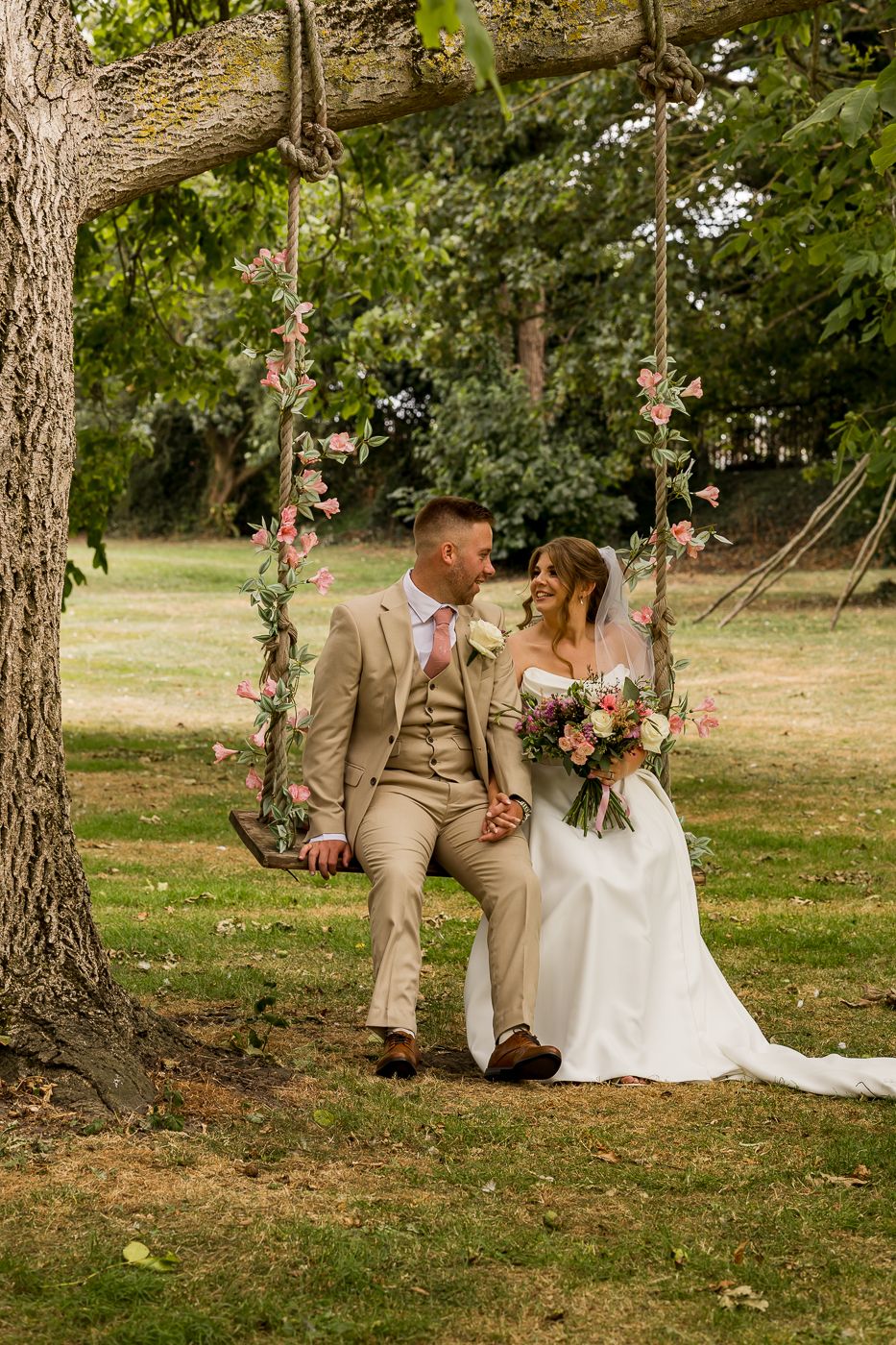 Bride and groom sit together on flower-adorned rope swing beneath large tree in lush garden setting.