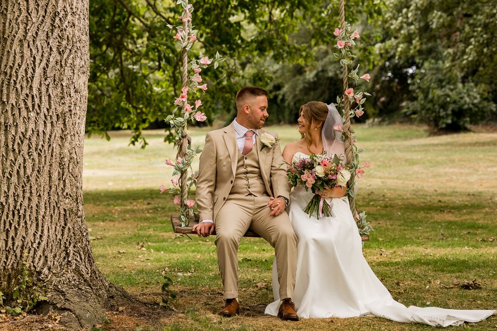 Bride and groom sitting on a floral swing under a tree in a lush garden on their wedding day.