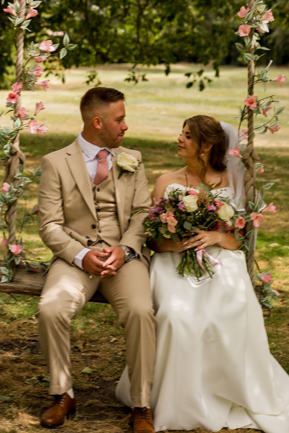 A bride and groom sit on a floral swing surrounded by pink flowers and greenery, smiling at each other on their wedding day.
