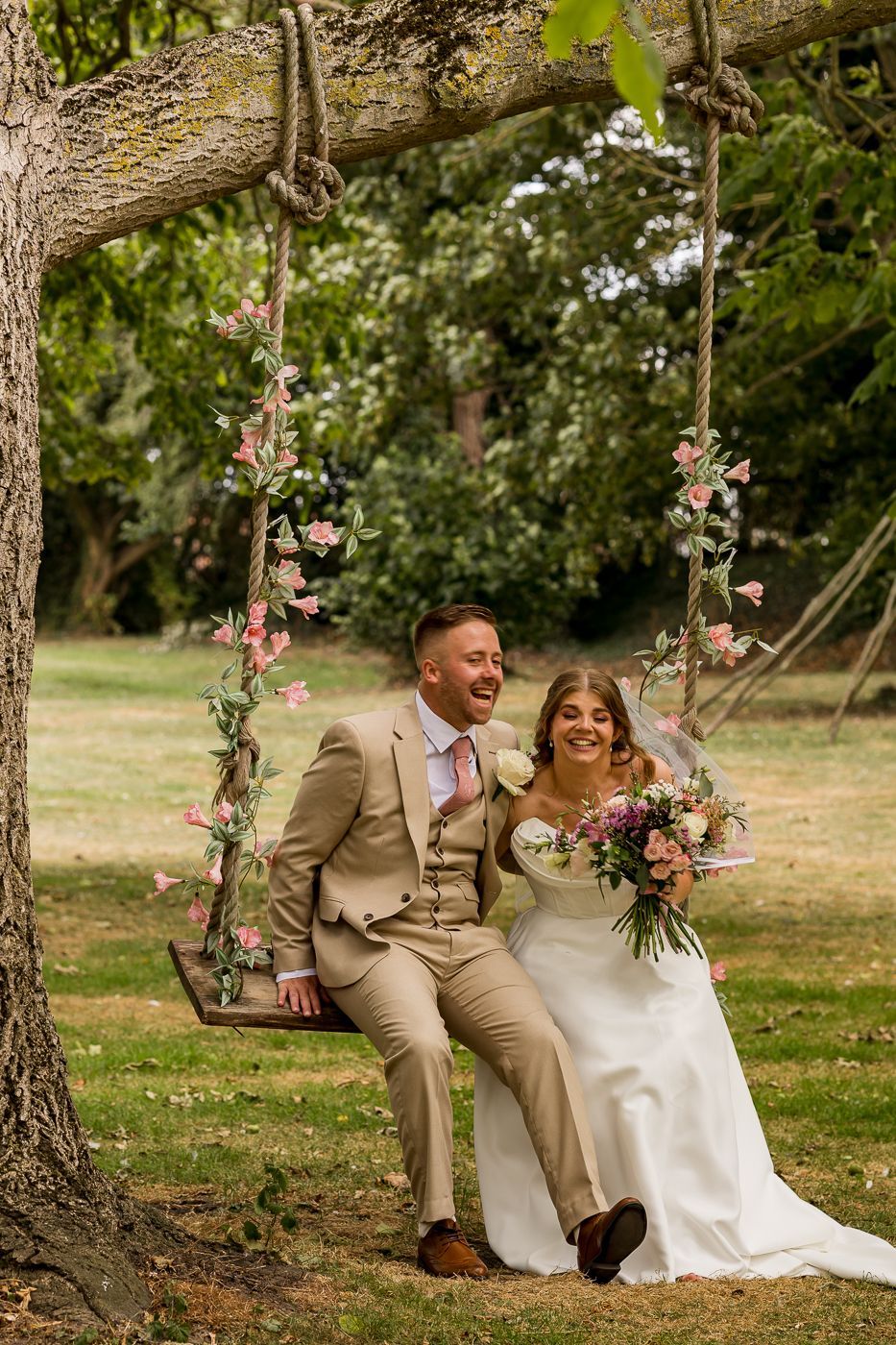 Bride and groom sitting together on a floral decorated rope swing in a lush green garden on their wedding day.