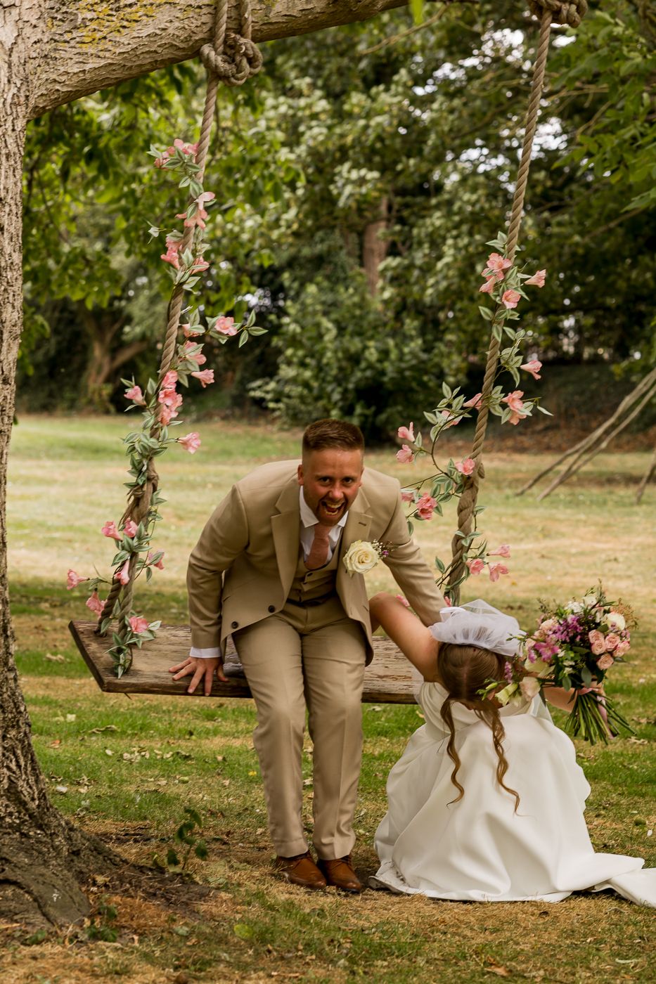 Groom in tan suit on floral swing laughing with bride in white dress holding bouquet outdoors.