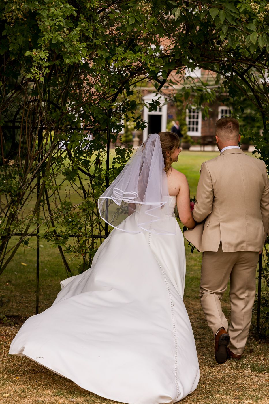 Bride in white gown and veil walks with groom in tan suit through garden arch at outdoor wedding.