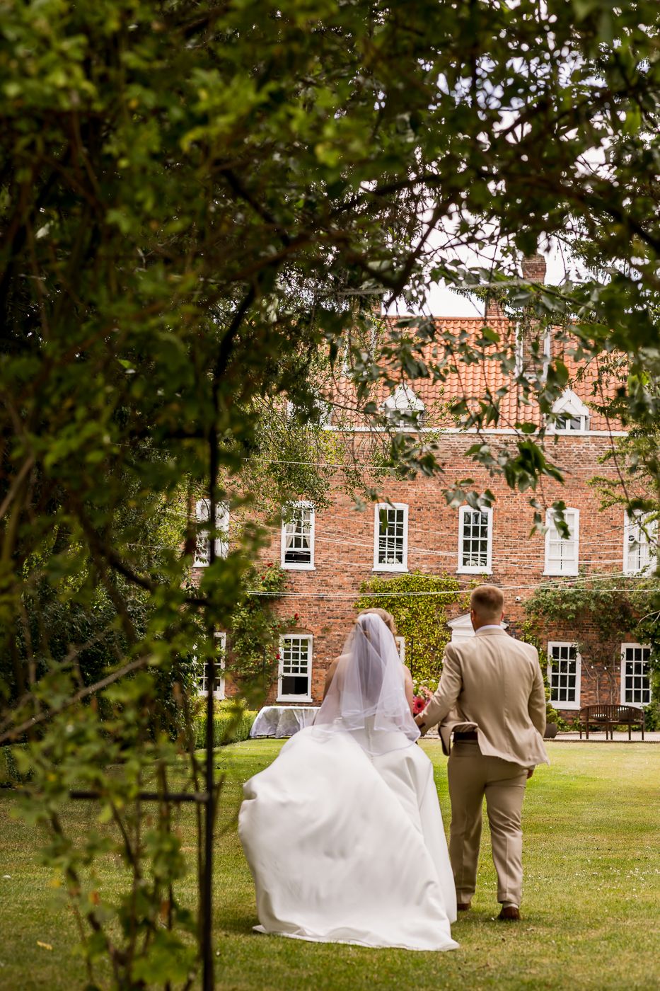Bride and groom walk hand in hand across lush garden toward elegant red brick wedding venue.