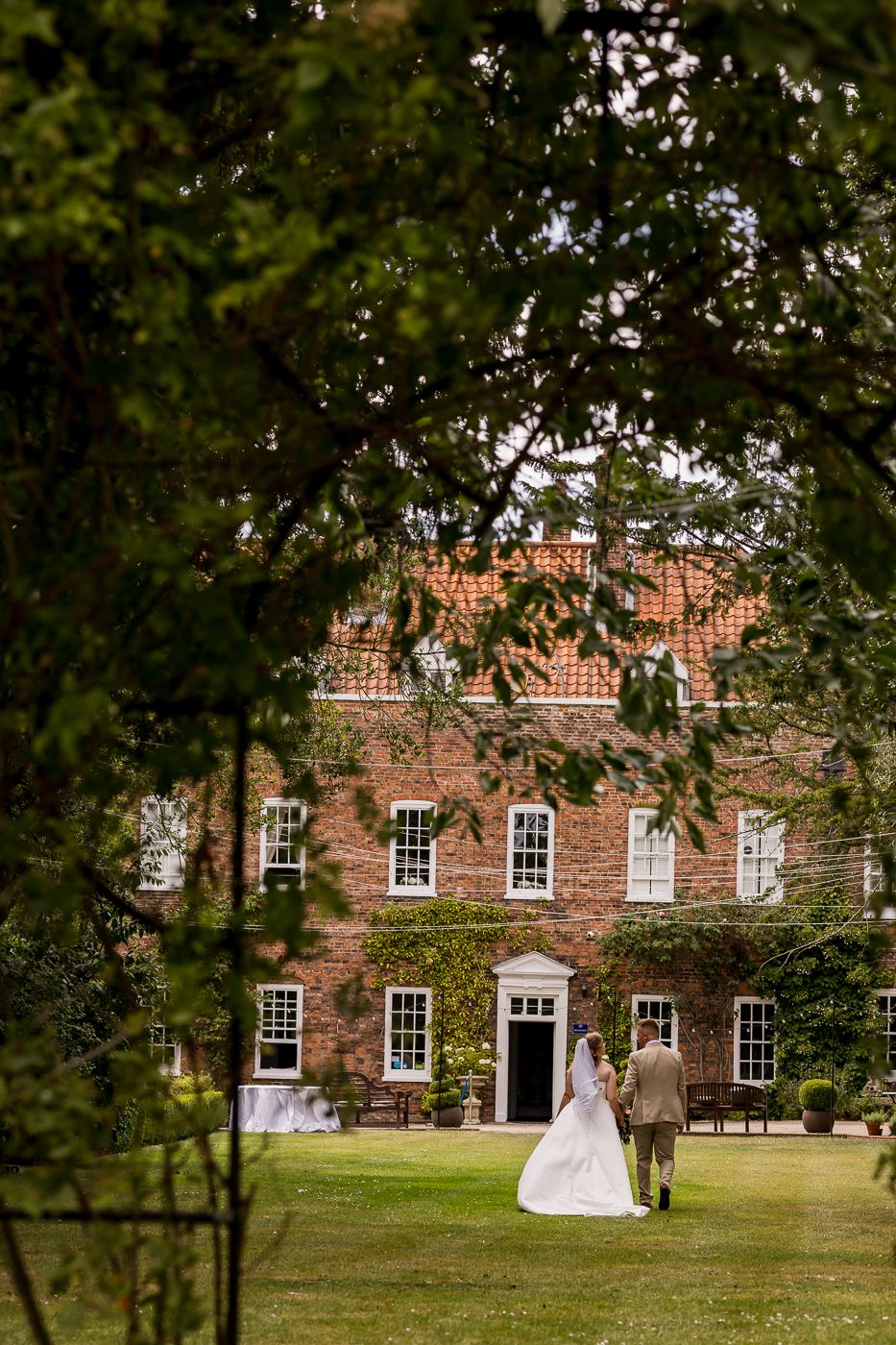 Bride and groom walking hand in hand toward an elegant ivy-covered red brick country house wedding venue.