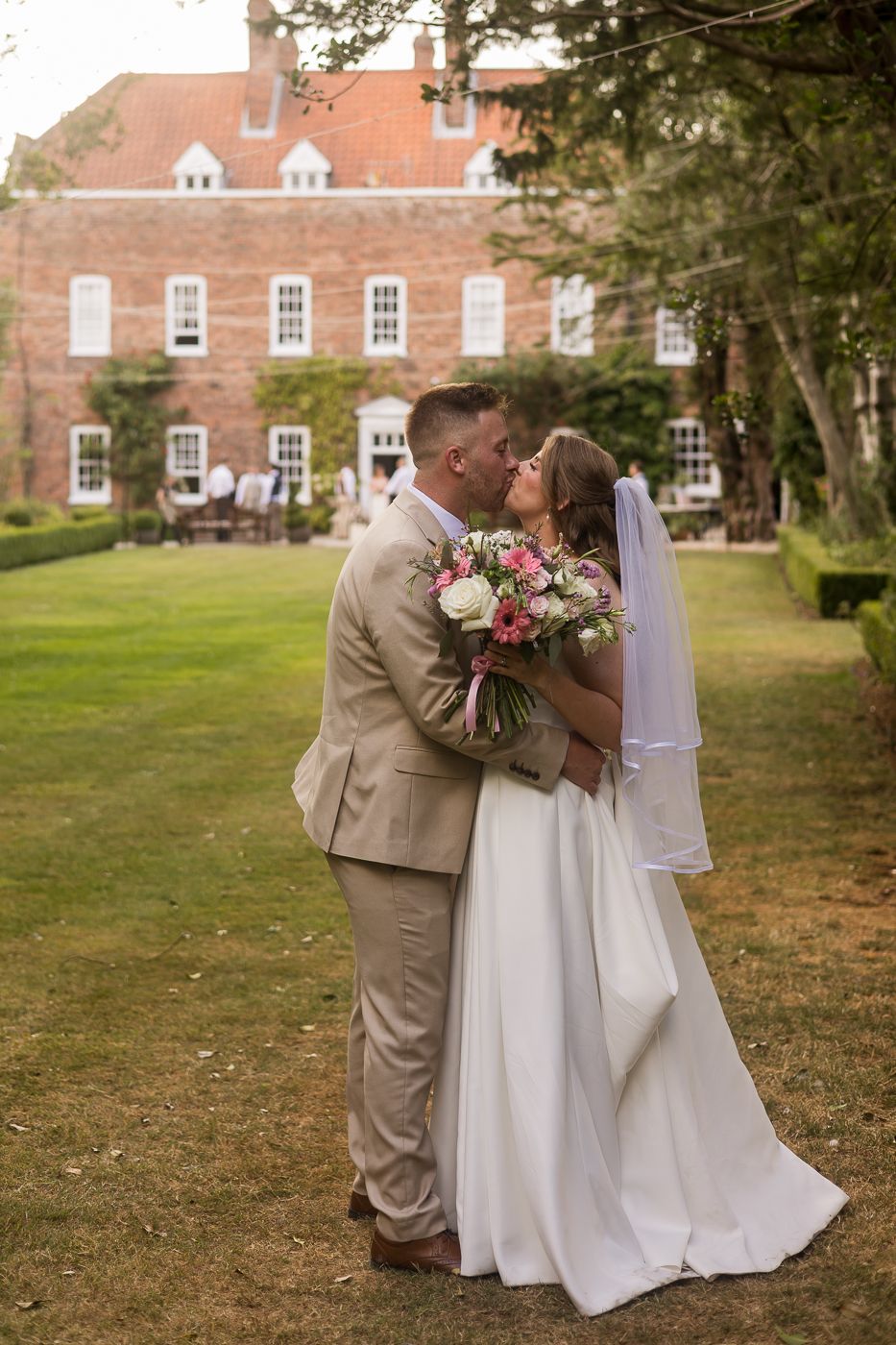 Bride and groom share a romantic kiss on lush garden grounds of elegant brick manor house on their wedding day.