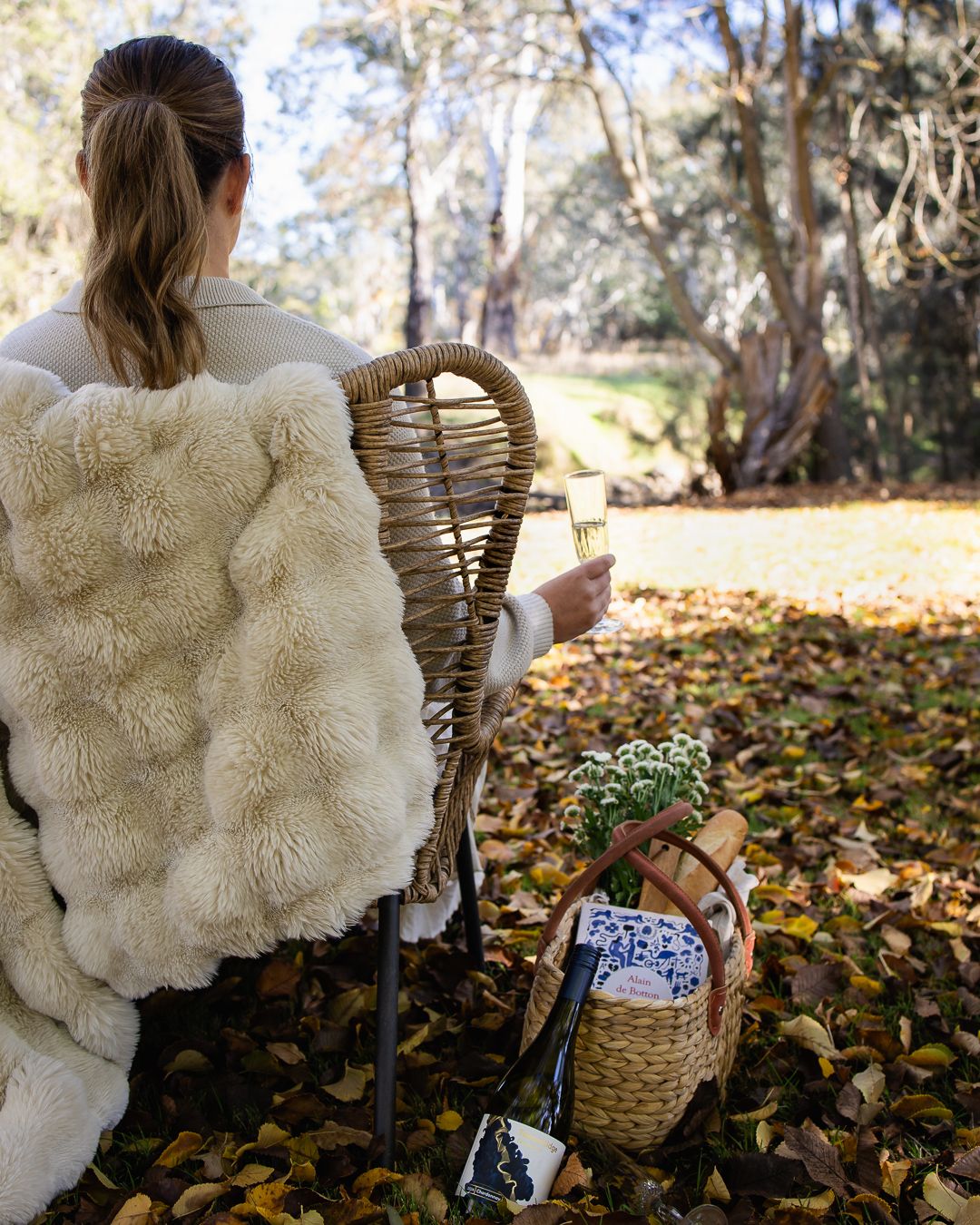 Woman relaxing in wicker chair outdoors with fluffy cream blanket, holding wine glass, beside woven basket in autumn leaves.