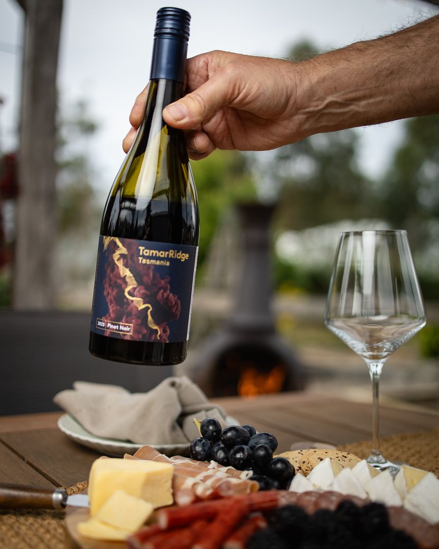 Hand holding Tamar Ridge Tasmania Pinot Noir wine bottle over a rustic charcuterie board with cheese, grapes, and an empty wine glass.