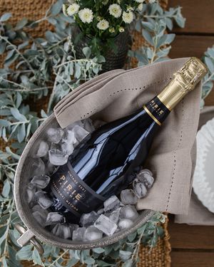Pirée sparkling wine bottle chilling on ice in a linen-lined bowl, surrounded by eucalyptus and white flowers.