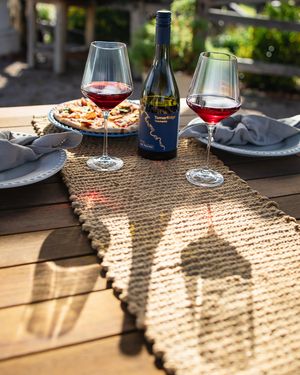 Red wine bottle and two glasses on rustic table with pizza, burlap runner, and warm sunlight casting shadows outdoors.