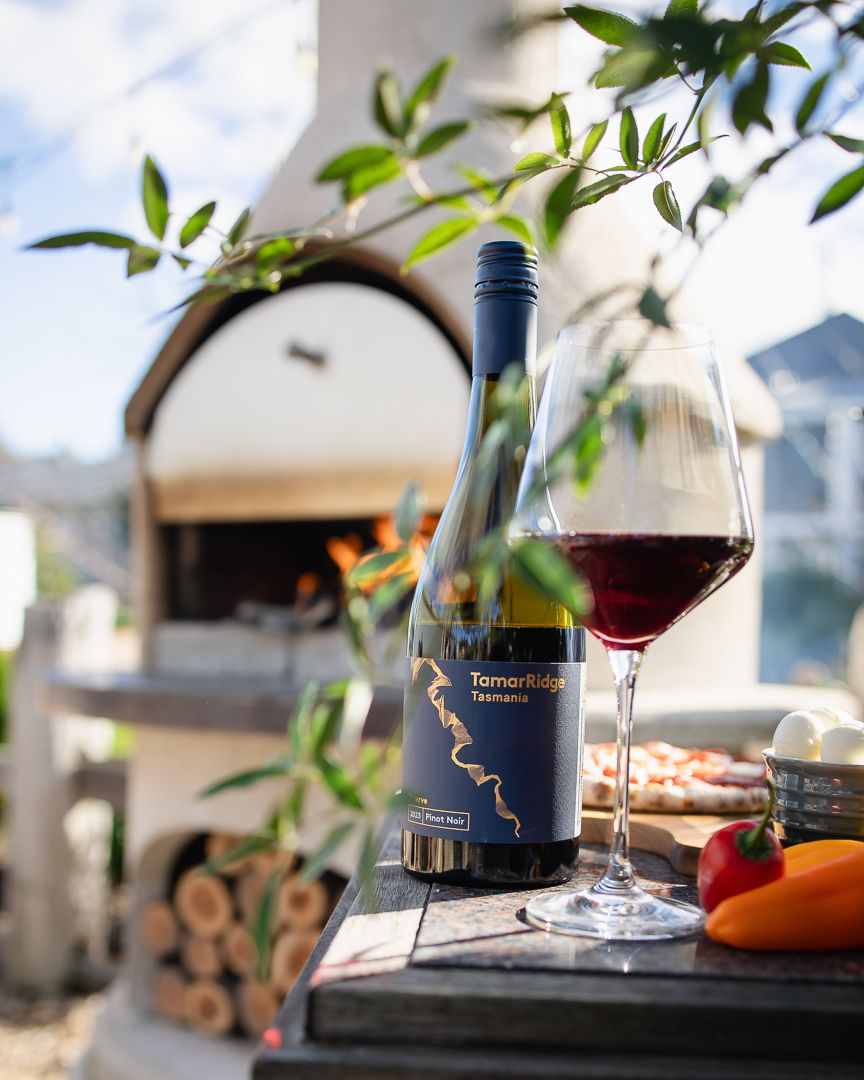 Tamar Ridge Tasmania Pinot Noir bottle and glass beside wood-fired oven with fresh vegetables outdoors.