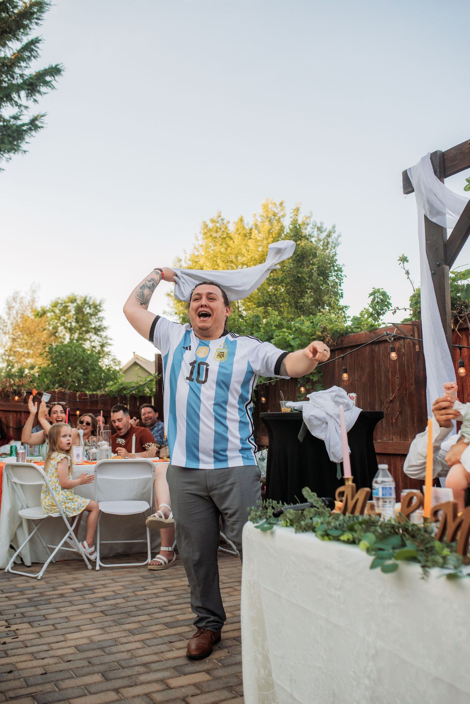 Man in Argentina soccer jersey celebrating outdoors at a backyard party, raising shirt above head in excitement.
