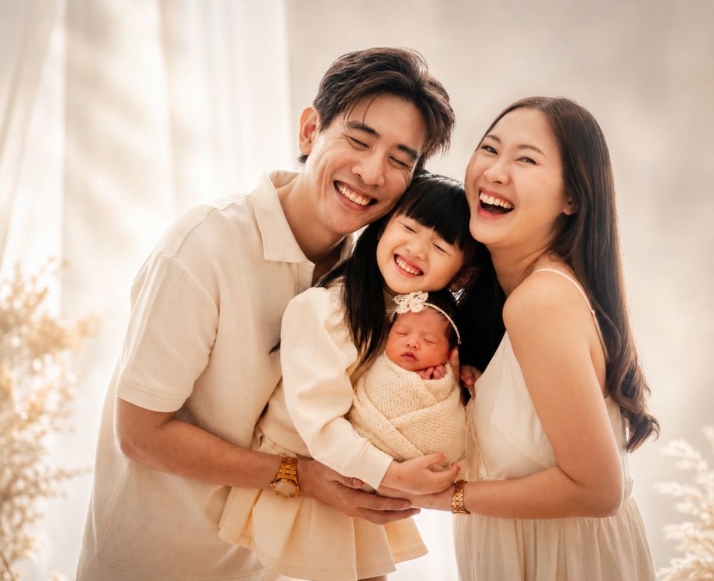 Happy Asian family of four smiling together, parents holding toddler girl and swaddled newborn baby in a bright studio.
