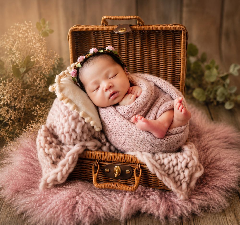 Newborn baby sleeping peacefully in a wicker suitcase, wrapped in pink knit blanket with floral headband.