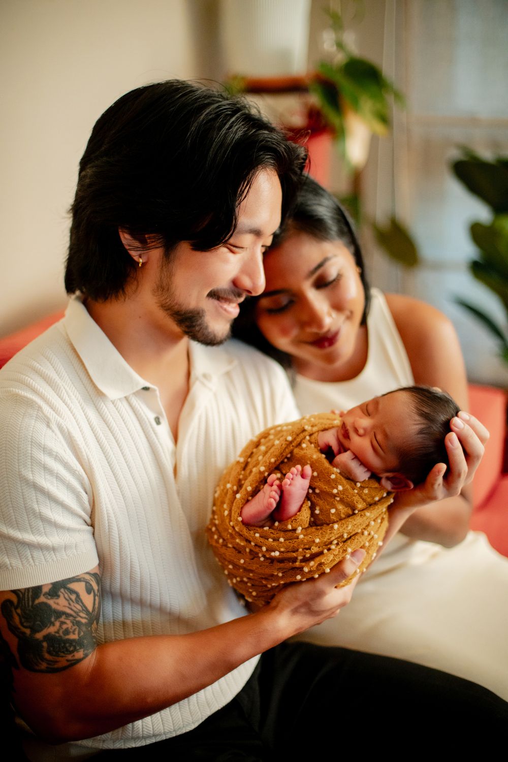 Young couple smiling and holding their sleeping newborn baby wrapped in a golden blanket at home.