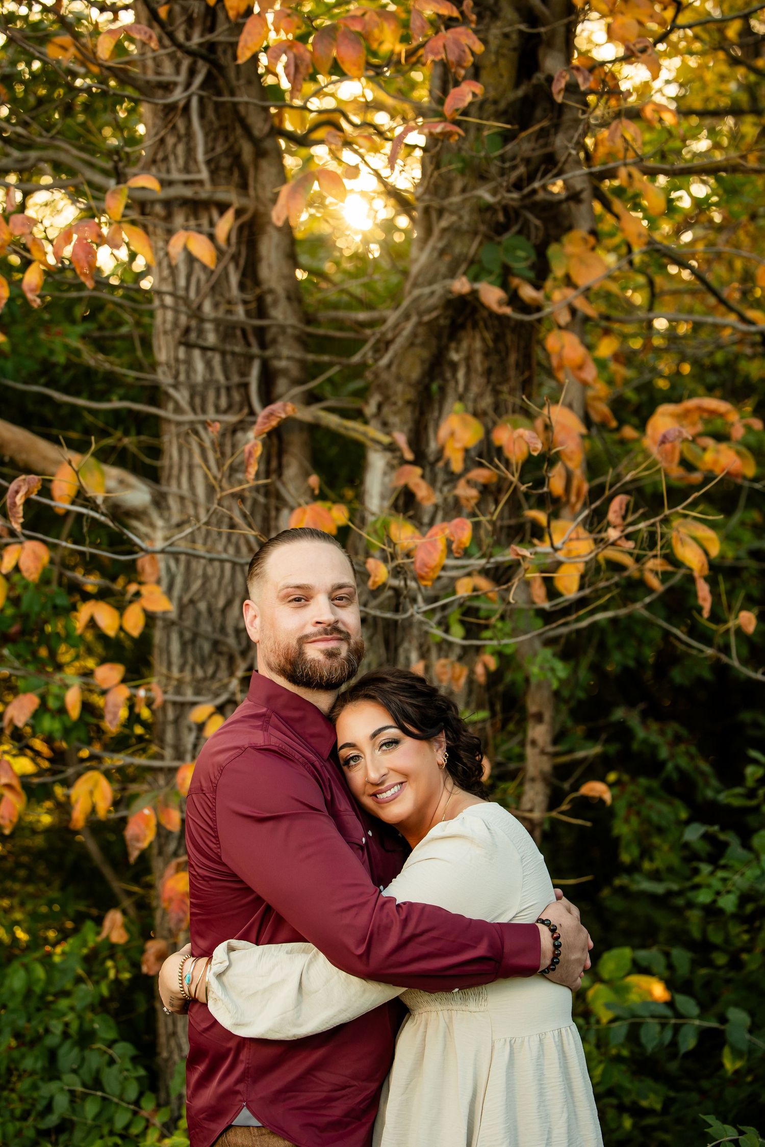 A couple embraces warmly among autumn foliage trees at golden hour, the woman smiling brightly in a cream dress.