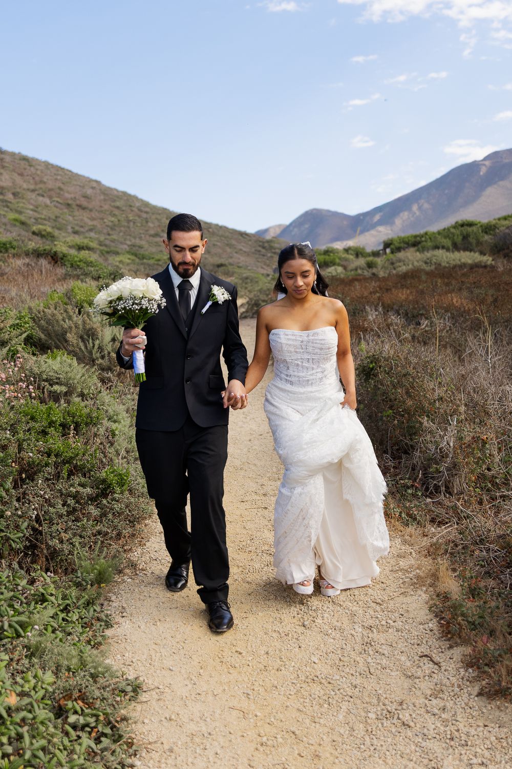A bride and groom walk hand-in-hand along a scenic mountain trail, she in a white gown, he in a black suit holding flowers.