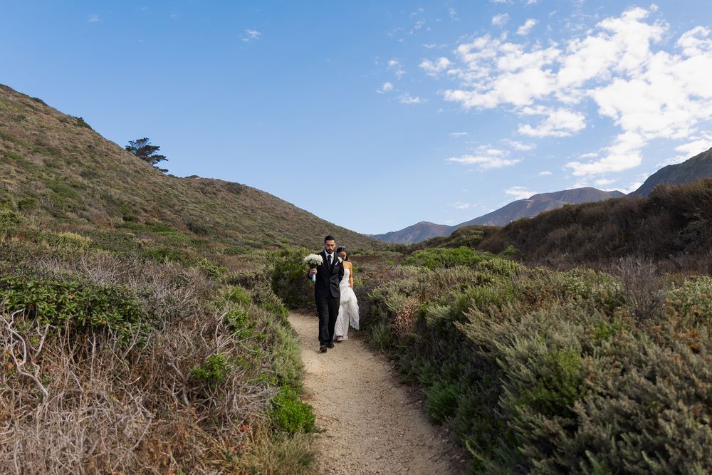A couple in wedding attire walks a scenic coastal trail surrounded by green hills and mountains under a blue sky.