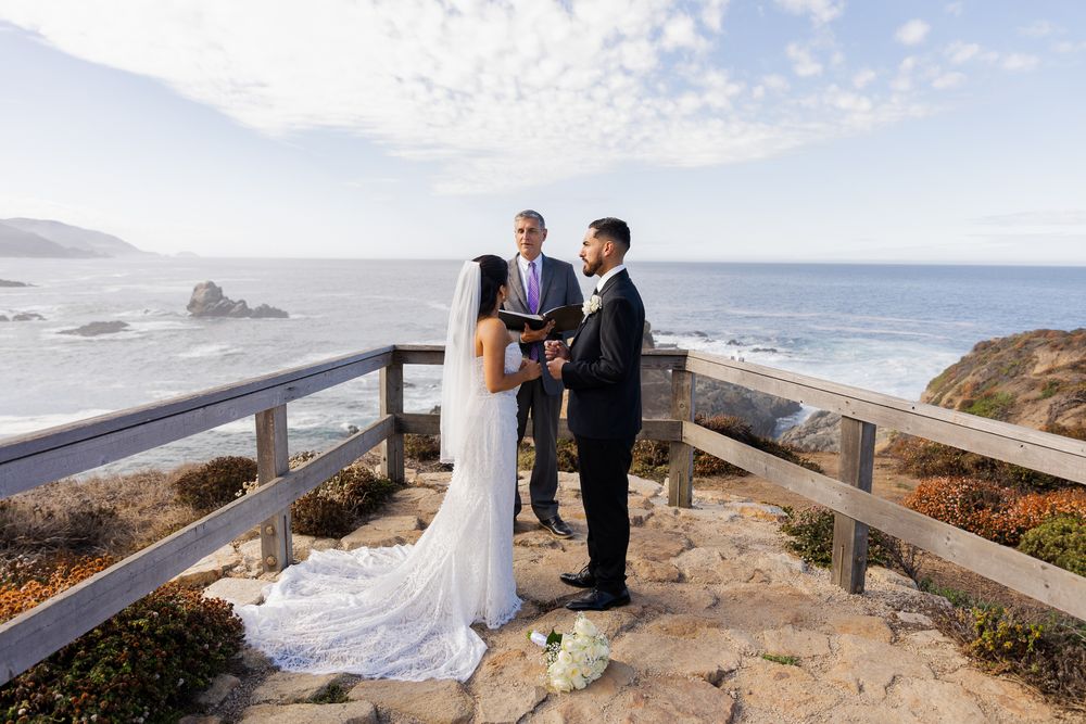 Couple exchanges vows at scenic coastal overlook with ocean views, officiant holding book, wooden railing, rocky shoreline backdrop.