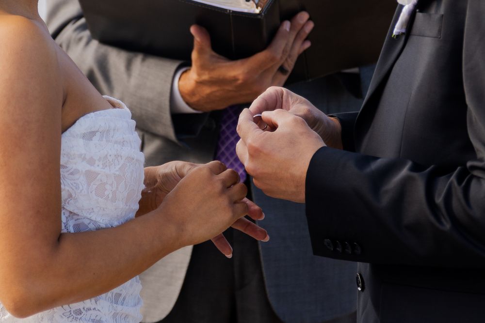 Bride and groom exchanging rings during wedding ceremony with officiant holding book in background.
